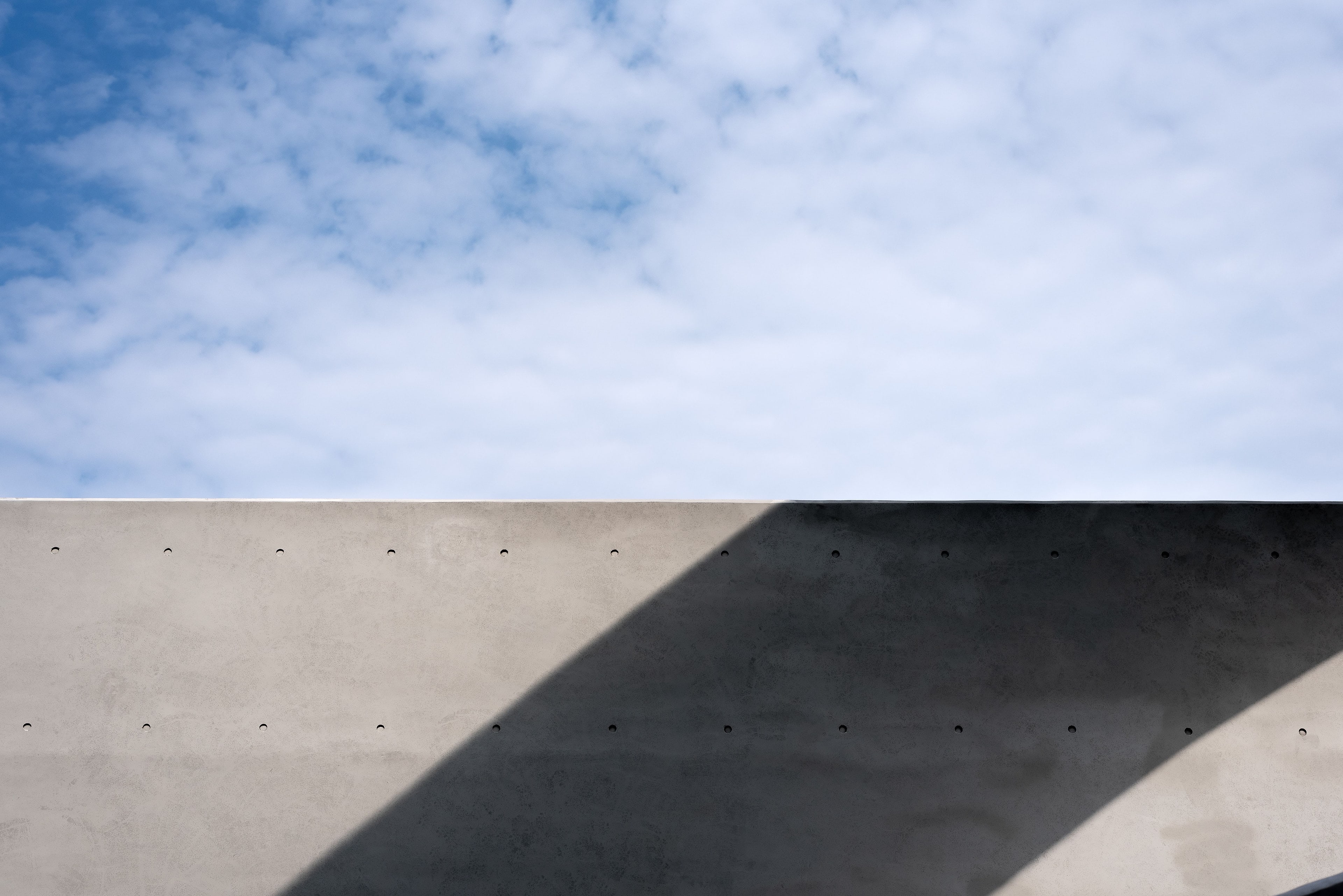Mur de béton nu et son ombre portée, sous un ciel bleu et nuageux.