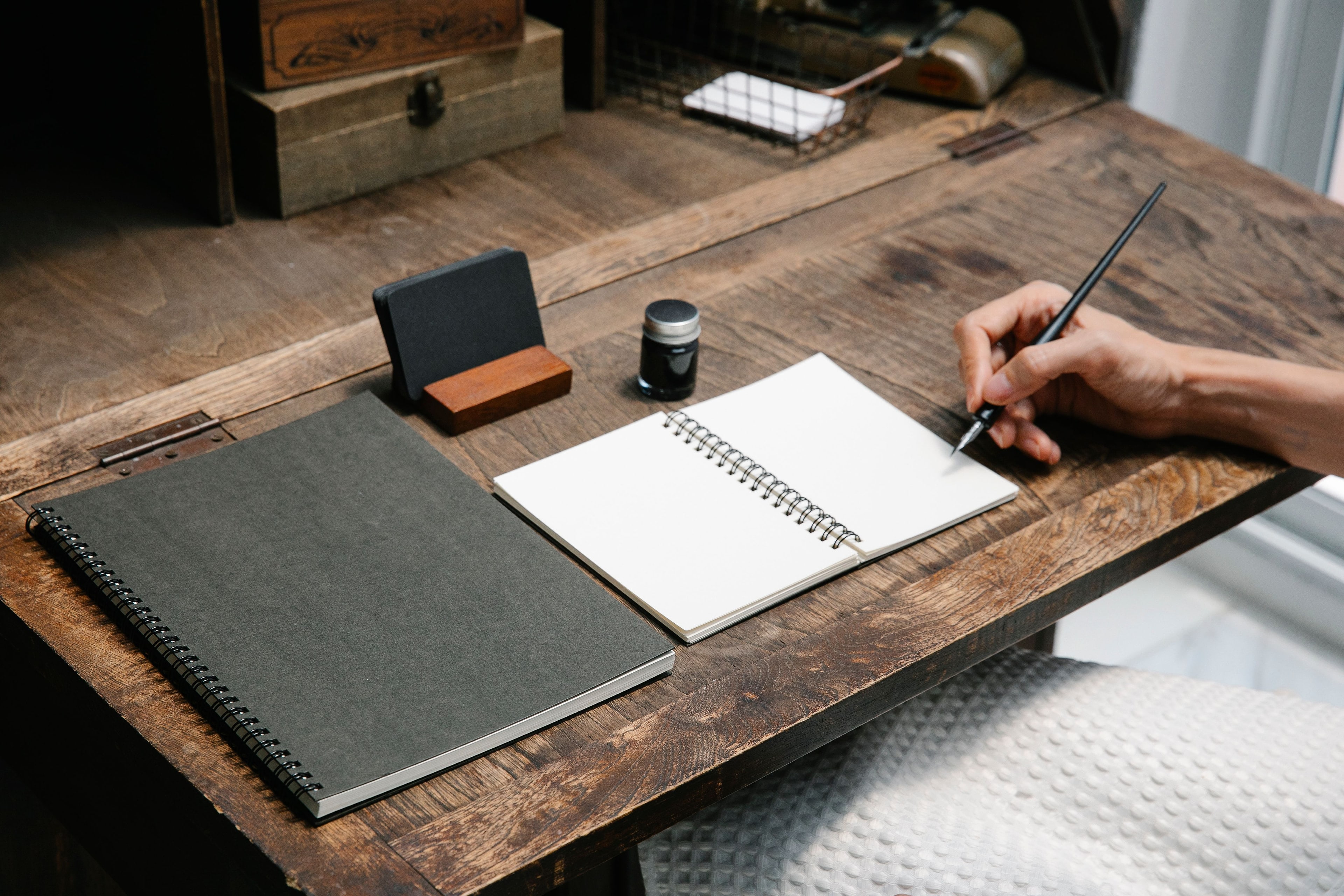 Une main écrit dans un carnet ouvert sur un bureau en bois.