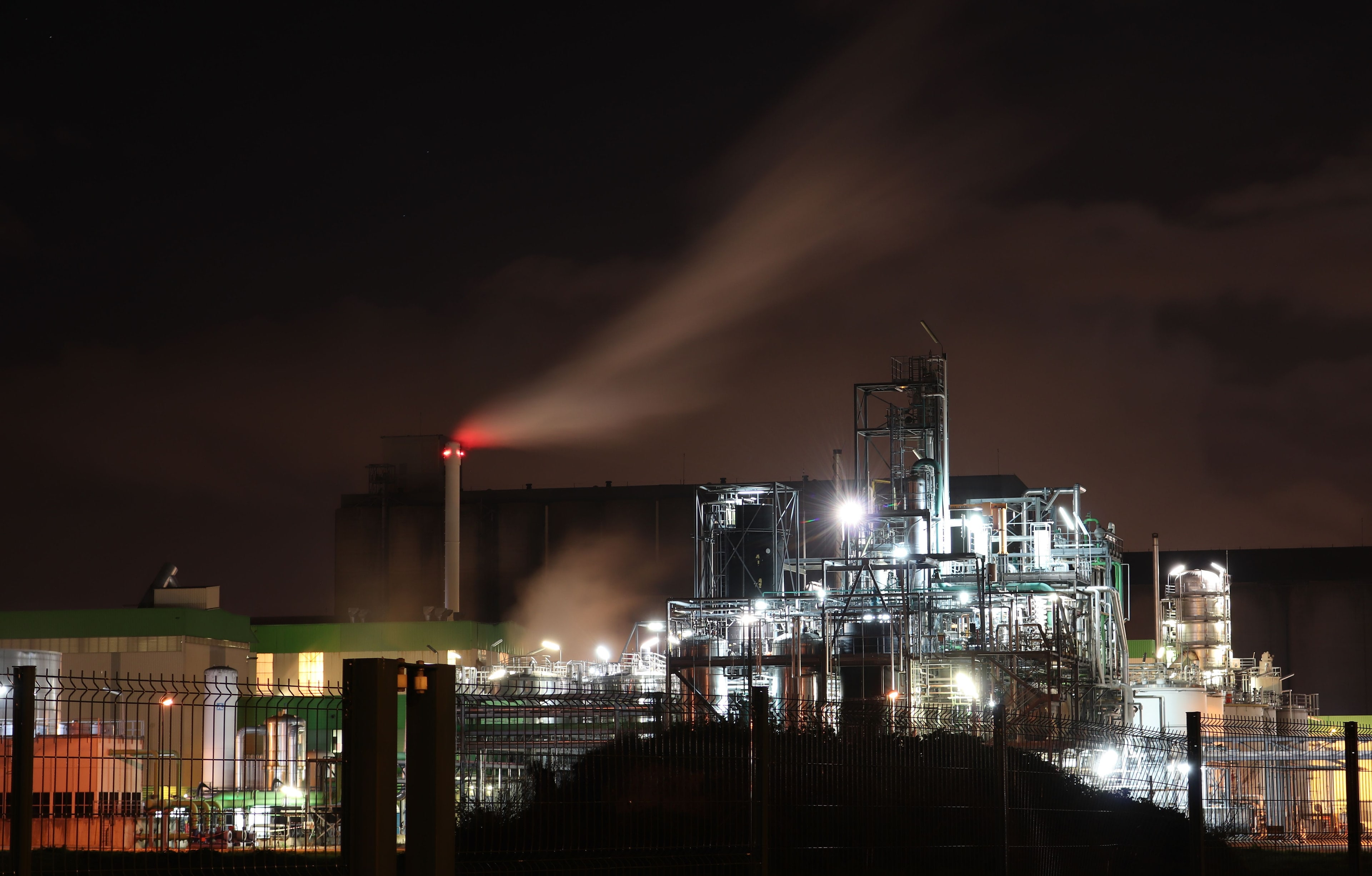 Raffinerie illuminée la nuit, un panache de fumée au-dessus des installations.